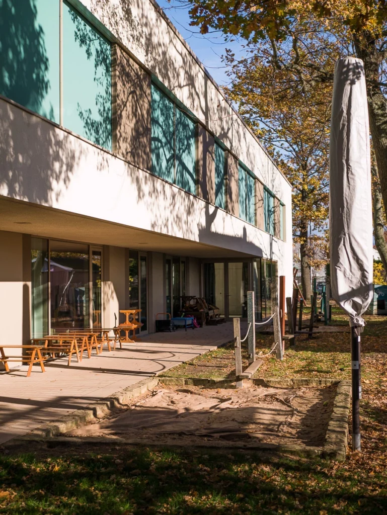Gebäude mit grünen Fenstern und Terrasse, leere Holzbank, Sandkasten im Vordergrund, herbstliche Bäume im Hintergrund.