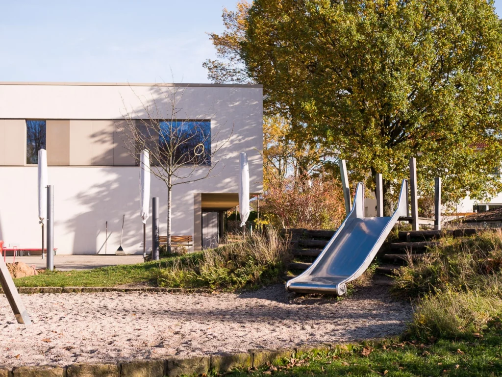 Spielplatz mit Rutsche vor einem modernen, zweistöckigen Gebäude, umgeben von Bäumen und Rasenfläche.