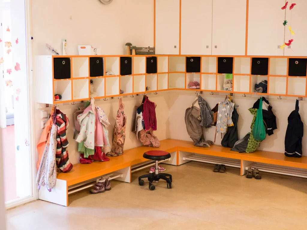 Garderobe in einem Kindergarten mit Haken, Fächern und bunter Kinderkleidung. Ein Drehhocker steht im Raum.
