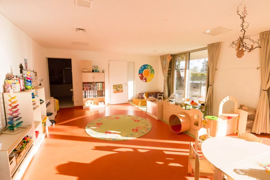 Heller Kindergartenraum mit Spielzeug, runden Teppichen, Regalen, Glastüren und Außenausblick.