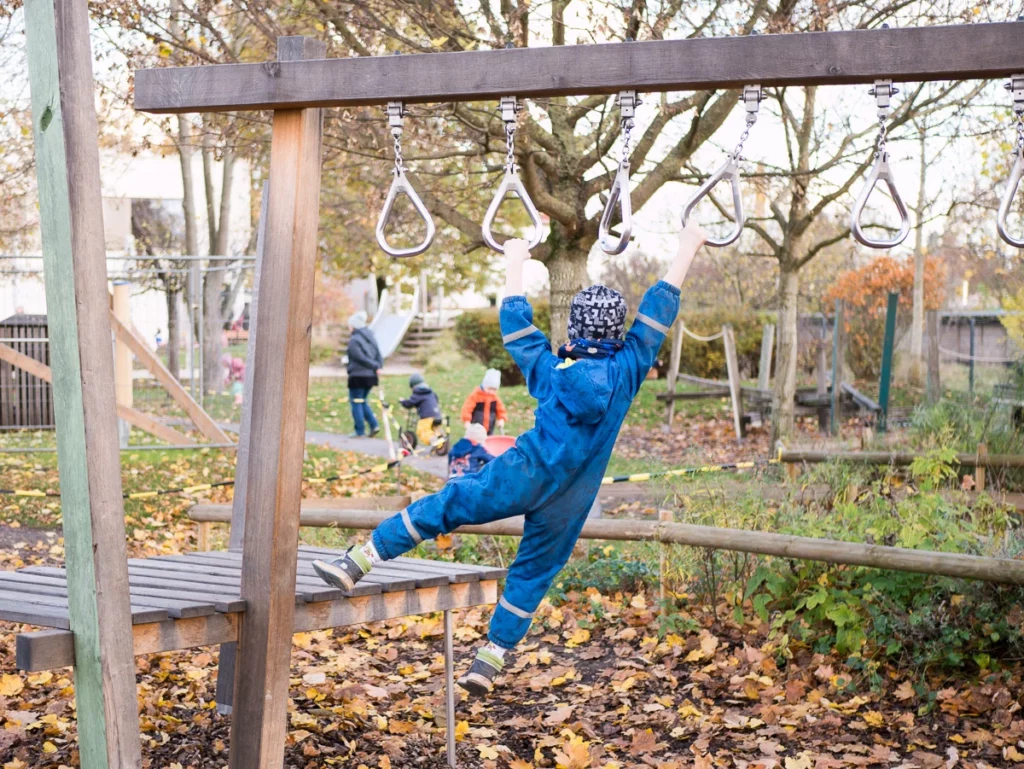 Kind auf Spielplatz klettert an Hangelringen, Herbstlaub am Boden, Bäume im Hintergrund. Weitere Kinder und Erwachsene spielen.