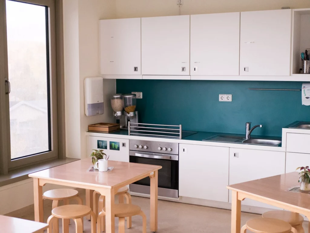 Moderne Küche mit weißen Schränken, Herd, Spüle, hellem Holztisch, Hockern und großem Fenster.