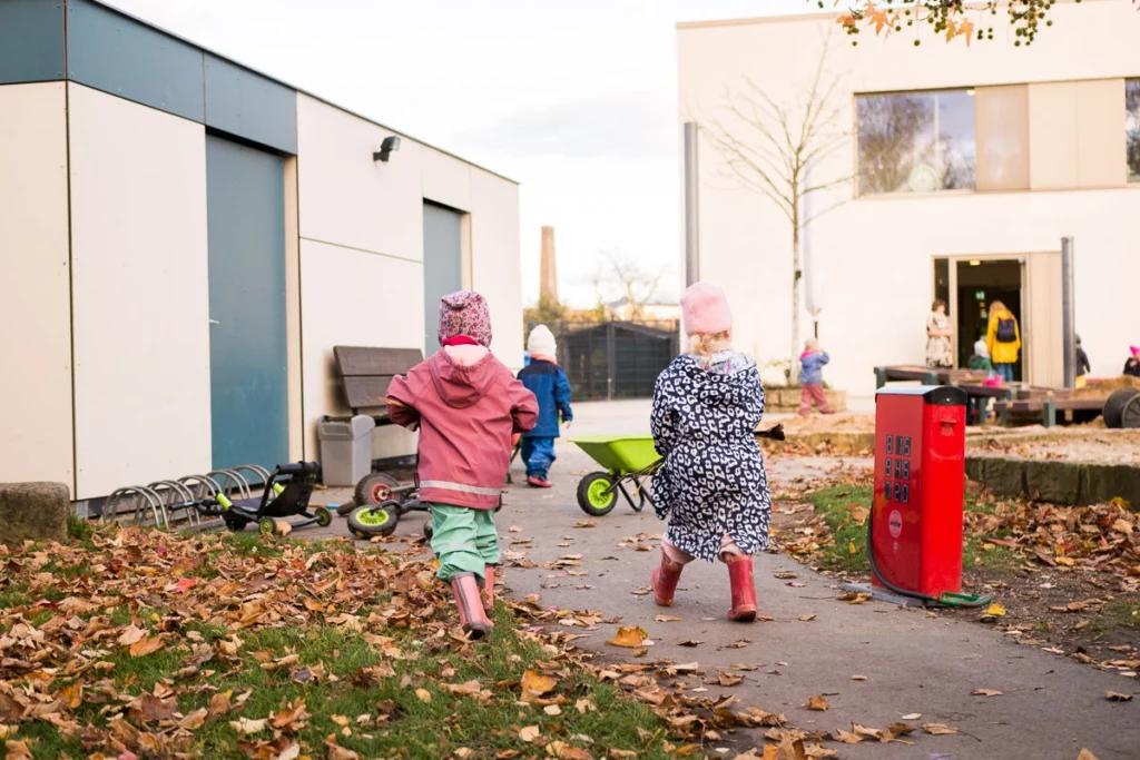 Kinder spielen im Freien zwischen Gebäuden, mit Laub bedecktem Boden und einem kleinen grünen Handkarren.